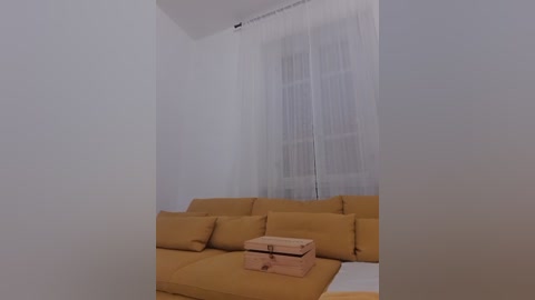 Video of a minimalist living room with a mustard-yellow sectional sofa, sheer white curtains, and a wooden storage box on a white floor. The room's walls are plain white.