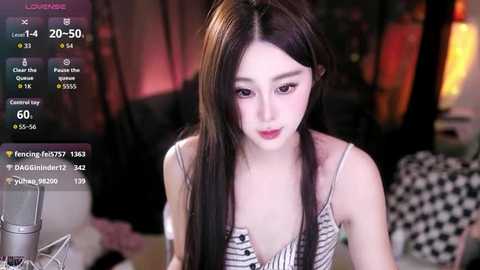 A video of a young Asian woman with long black hair, fair skin, and large eyes, wearing a striped tank top, sitting in a dimly lit room with a microphone in front of her.