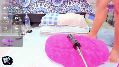 Video of a bedroom with a heart-shaped pink cushion and a hand using a vibrator, surrounded by a blue and white mandala tapestry and pillows.