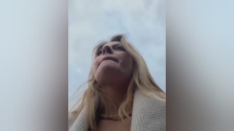 Video of a Caucasian woman with long, wavy blonde hair and fair skin, wearing a beige sweater, looking upward with a serene expression. The background is a blurred, light-colored sky.