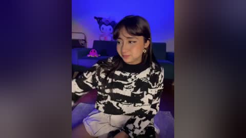 A video of a young Asian woman with long black hair, wearing a black-and-white patterned sweater and white shorts, sitting on a carpeted floor, in a dimly lit room with a blue light and a cartoon character poster on the wall.