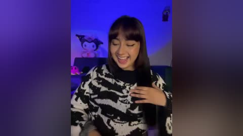 A video of a young Asian woman with straight black hair, smiling and holding a phone, wearing a black and white patterned sweater. Background includes a plush toy and a blue-lit room.