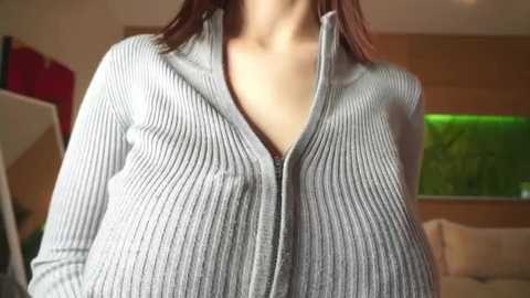 Video of a woman in a light gray, ribbed zip-up sweater, revealing cleavage. Background shows a modern living room with wooden furniture, a green fish tank, and a white couch.