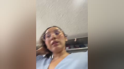 A blurred video of a woman with curly hair and glasses, wearing a light blue shirt, standing indoors with a textured white ceiling and bookshelves in the background.