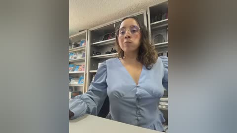 A video of a middle-aged woman with medium-length brown hair, wearing glasses and a light blue blouse, sitting behind a desk in an office with bookshelves and medical supplies in the background.