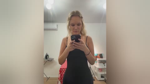 Video of a blonde woman in a black tank top taking a mirror selfie in a bright room with white walls, wooden floor, and an air conditioner.