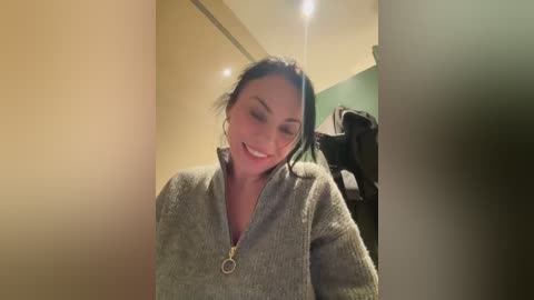 Video of a smiling, light-skinned woman with dark hair, wearing a gray zip-up sweater, standing in a hallway with beige walls and green accents, and a blurred person in the background.