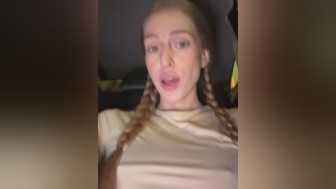 Video of a young woman with fair skin, blonde braided hair, and light beige shirt, making an open-mouthed expression, blurred background.