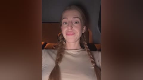 A video of a young woman with light skin and braided blonde hair, smiling broadly, wearing a beige top, seated in a dimly lit car interior.
