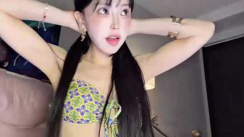 Video of an East Asian woman with long black hair, wearing a floral-patterned bikini top, posing with arms raised, in a dimly lit room with dark furniture.