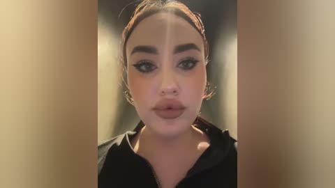 Video of a young woman with light brown skin, dark hair, and bold makeup, including black eyeliner and lipstick. She wears a black jacket. The background is blurred, emphasizing her face.