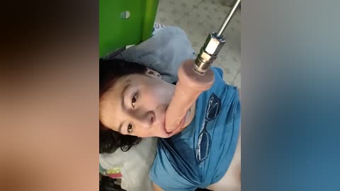 A video shows a young Asian woman with black hair, wearing a blue shirt, performing oral sex on a large, erect penis. The background includes a green chair and beige carpet.