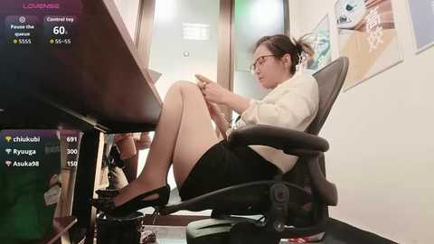 Video of a young East Asian woman with glasses and short black hair, wearing a white blouse and black skirt, seated at a desk in a modern office, playing a video game.