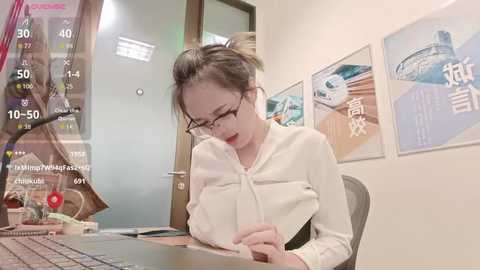 Video of an Asian woman with glasses, wearing a white blouse, typing on a computer in an office with white walls and framed posters.