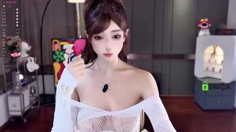 Video of a young Asian woman with fair skin, large eyes, and a slim figure, wearing a white, off-the-shoulder top. She sits at a desk with colorful artwork and a sewing machine in the background.