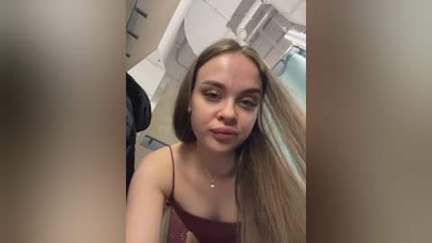 Video of a young woman with long, straight, light brown hair, wearing a maroon spaghetti-strap top, standing in an airplane aisle, looking forward.