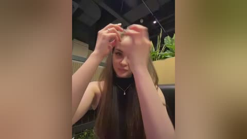 Video of a young woman with long brown hair, using her hands to comb through it. She wears a black turtleneck and has fair skin. Indoor setting with plants in the background.