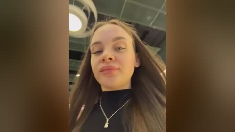 Video of a young woman with straight, long brown hair and light skin, wearing a black turtleneck, standing indoors with a ceiling light and blurred background.