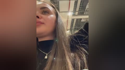 Media: A close-up video of a young woman with long, straight, light brown hair, wearing a black top and silver necklace, taken in an industrial setting with visible pipes and metal beams.