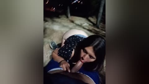Video of a dark-haired woman, wearing a black floral top, performing oral sex on a man lying on a dirt path at night.