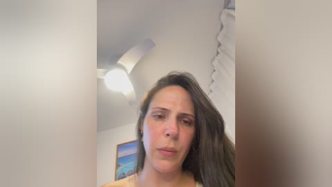 A video of a woman with long brown hair and light skin, wearing a yellow top, standing in a dimly lit room with a ceiling fan and a partially visible painting on the wall.