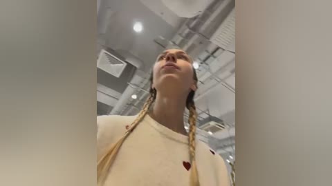 Video of a young woman with light skin and blonde braided hair, wearing a cream-colored sweater with a small red heart emblem, standing in a modern, industrial-style room with white walls and exposed pipes.