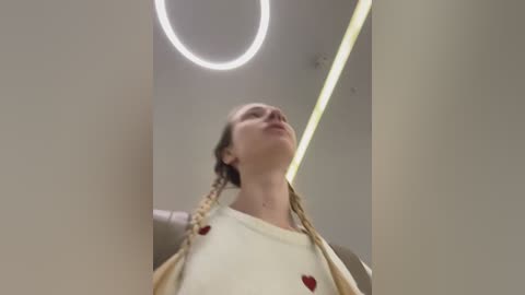 Video of a young woman with light skin and braided hair, wearing a white shirt with red buttons, standing in a modern, minimalist room with a circular ceiling light and beige walls.