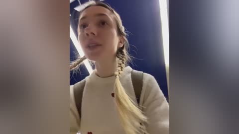 Video of a young Caucasian girl with fair skin, light brown hair in a braid, wearing a beige sweater and backpack, looking into the camera against a dark blue background.
