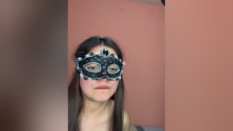 Video of a young woman with long brown hair, wearing an ornate black lace masquerade mask, set against a plain, muted orange wall. Her expression is neutral.