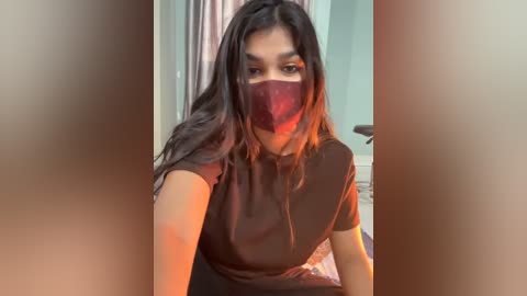 Video of a young woman with long, dark hair and medium skin tone, wearing a red face mask and a brown top, seated indoors with a blurred background.