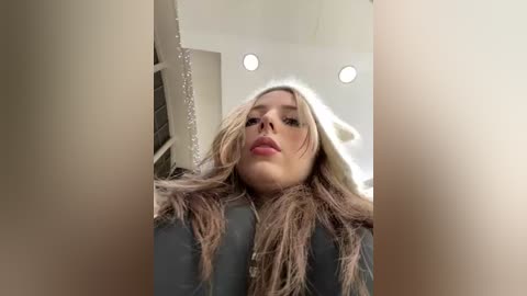 Video of a young Caucasian woman with long, wavy, platinum blonde hair, wearing a gray jacket, taken from a low angle. Background includes a white room with recessed lights.