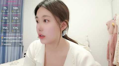 Video of an East Asian woman with light skin, brown hair tied back, wearing a white robe, in a bathroom with a shower curtain and hanging towels.