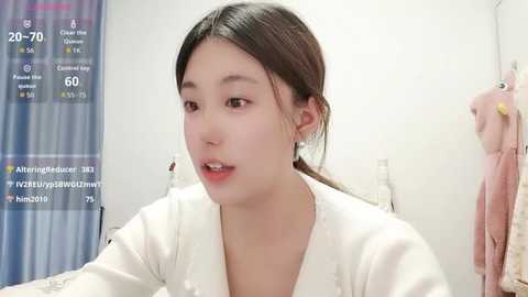 Video of an Asian woman with light skin, brown hair tied back, wearing a white robe, speaking in a bathroom with a pink towel hanging.