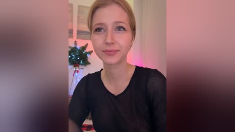 Video of a young, light-skinned woman with blonde hair, wearing a black mesh top, smiling slightly, in a room with festive decorations and soft pink lighting.