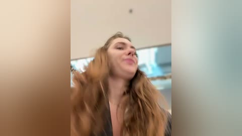 A blurry video of a woman with long, wavy hair, wearing a gray jacket, standing indoors with a blurred background featuring a beige wall and a hint of a window.