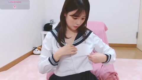 Video of an East Asian woman with long black hair, wearing a white sailor school uniform, adjusting her blouse in a pink room with a doll and a white door.