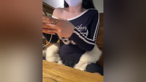 Video of a woman in a navy sweater with white trim, wearing a mask, holding a phone, seated on a wooden chair, with a blurred background.