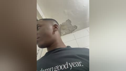 Video of a young Black man with short hair wearing a black shirt, taken from a low angle, showing his side profile. The background features a ceiling with a water stain and a tiled wall.