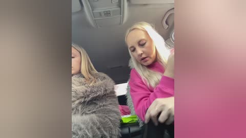 A video of a blonde woman in a pink sweater seated in a car, with another woman in a gray coat visible beside her. The image is slightly blurry.