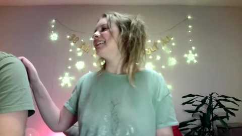 Media: Video of a young, smiling woman with messy blonde hair in a light green t-shirt, in a room with string lights and a potted plant.