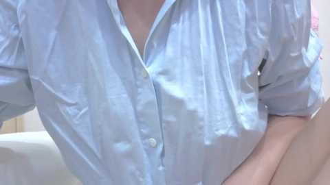 Video of a person in a light blue, button-up shirt with sleeves rolled up to the elbows. The background is blurred, showing a white couch.