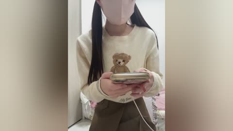 Video of a young woman with long black pigtails, wearing a beige sweater featuring a small teddy bear, pink mask, and brown skirt, holding a smartphone.