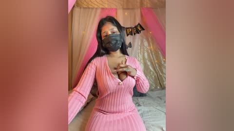 Video of a dark-skinned woman with long black hair, wearing a pink ribbed dress, black mask, and a black choker, in a cozy, pink-tinted room with \"PEA\" banner and fairy lights.