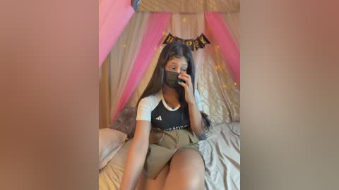 Video of a young woman with long black hair, wearing a black face mask, black top, and khaki shorts, sitting on a bed with beige sheets, surrounded by pink and gold tulle curtains and a \"Happy Birthday\" banner.