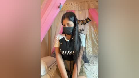 Media: Video of a young South Asian woman with long black hair, wearing a black and white \"Empire\" T-shirt and a black face mask, sitting on a bed with pink and white curtains.