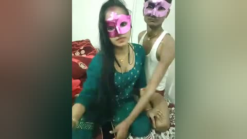 Video of two South Asian women in traditional outfits, wearing pink masquerade masks, sitting on a bed with red and white bedding.