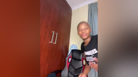 Video of a young Black man with short hair, wearing a black t-shirt, standing in a room with a wooden closet, a blue curtain, and a gaming chair.