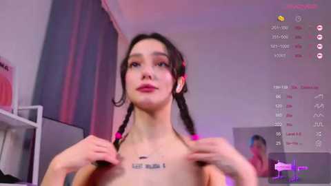 Video of a young woman with light skin, dark braided hair, and pink lipstick, topless, holding her breasts, in a dimly lit room with pink and purple lights, displaying her \"left\" tattoo.