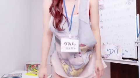 Video of a young woman with long, wavy red hair, wearing a sleeveless, light pink dress with a floral pattern, holding a lanyard. Background includes a whiteboard with drawings, a table with books, and a wall with colorful artwork.