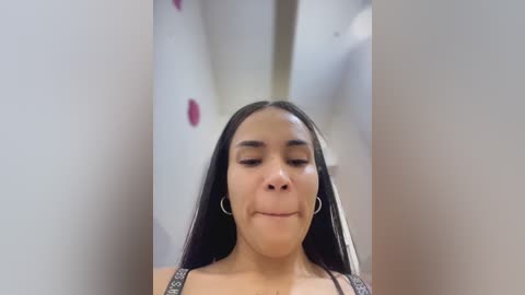 Video of a Latina woman with long black hair, wearing a black top, standing in a narrow hallway with white walls and pink dots.